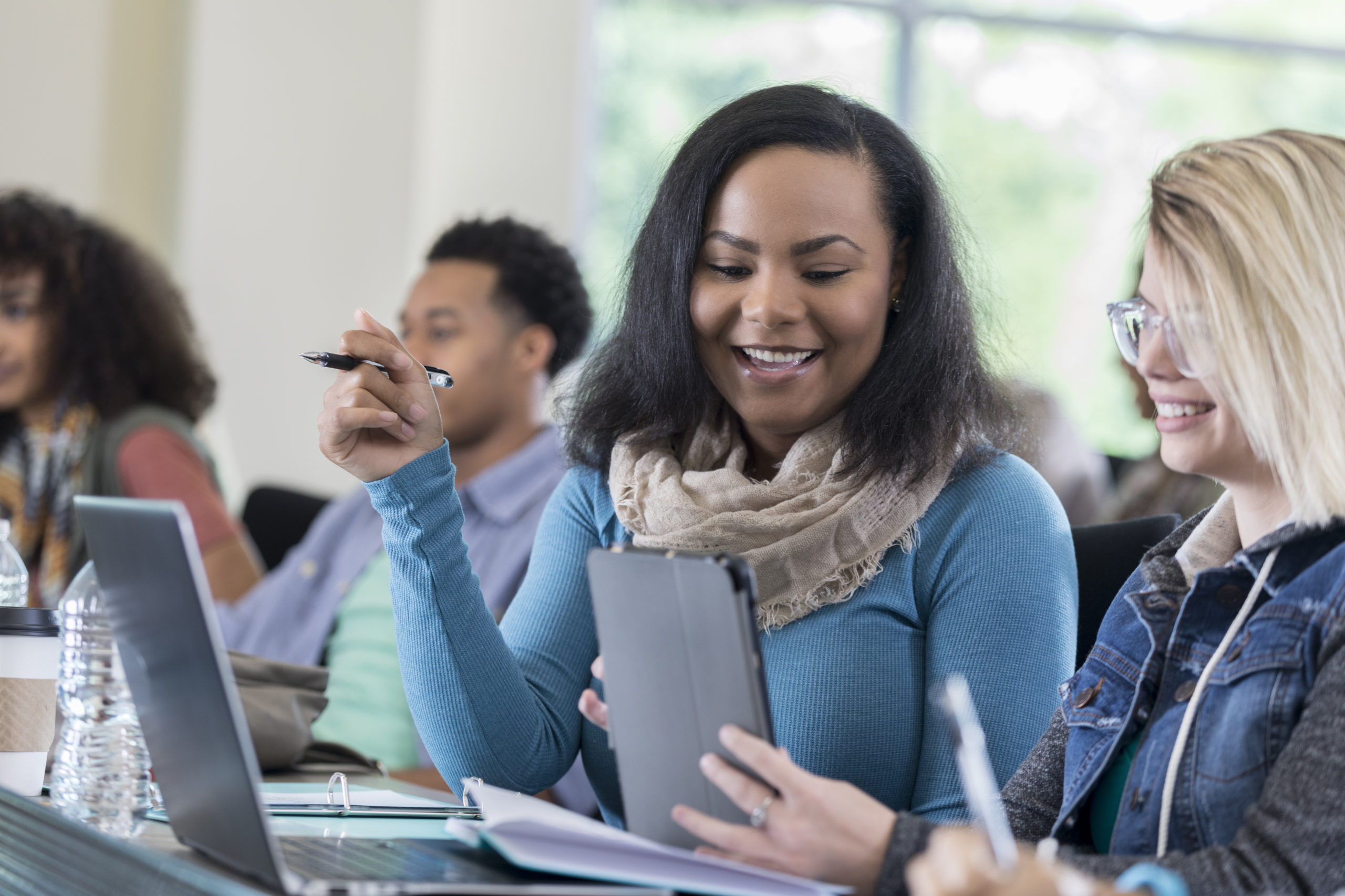 Pretty African American and Caucasian college friends use laptop to study before class. An open laptop is on the table in front of them. Students are in the background.